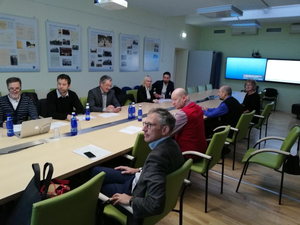 Grupa około dziesięciu osób uczestniczących w spotkaniu roboczym lub warsztatach w jasnej sali konferencyjnej. Uczestnicy siedzą przy długim, jasnym stole w zielonych fotelach, a na ścianie za nimi widoczne są plakaty informacyjne.