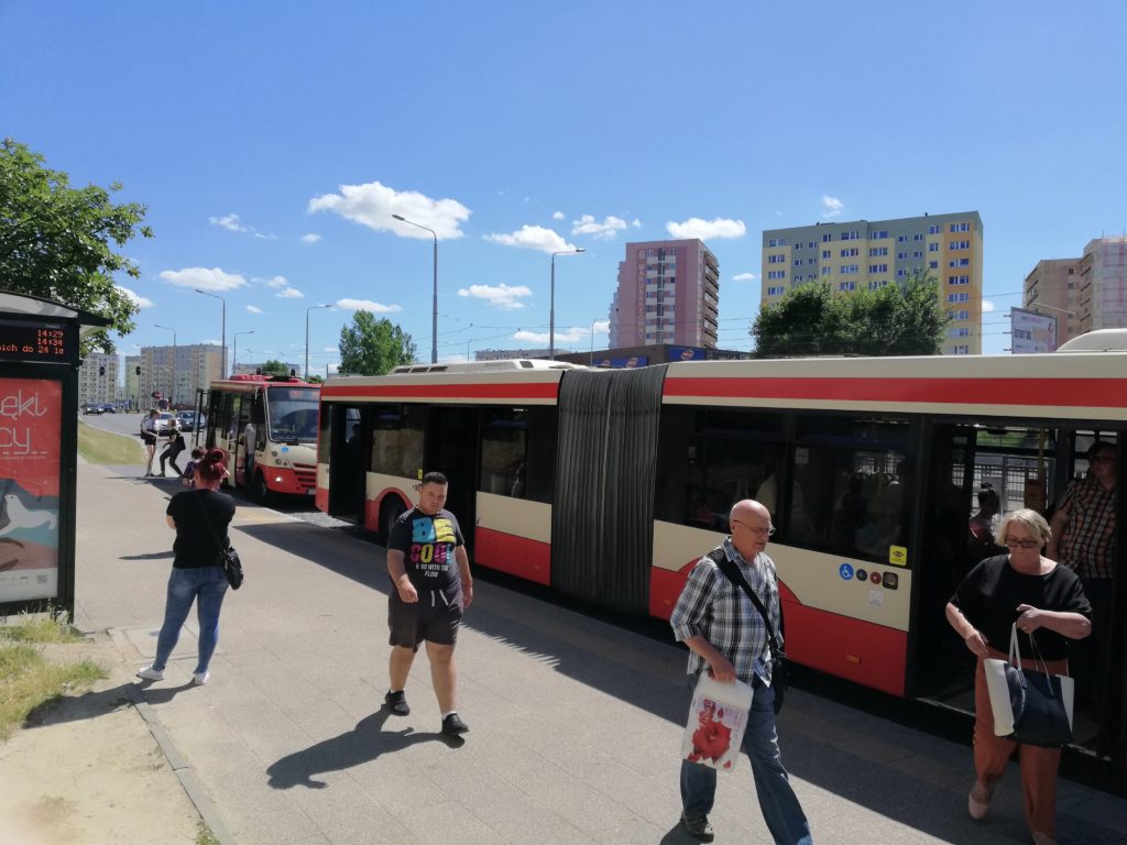 Pasażerowie na przystanku obok czerwono-białego autobusu przegubowego w słoneczny dzień