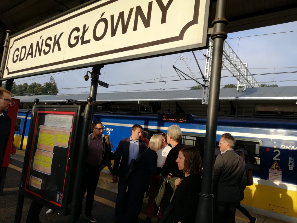 Grupa osób na peronie pod szyldem „Gdańsk Główny” obok pociągu SKM.